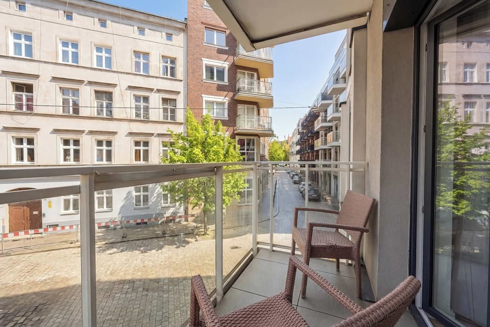A city-view balcony is furnished with a comfortable brown wicker chair, offering a perfect spot to relax. The glass railing provides an unobstructed view of the surrounding historic architecture.