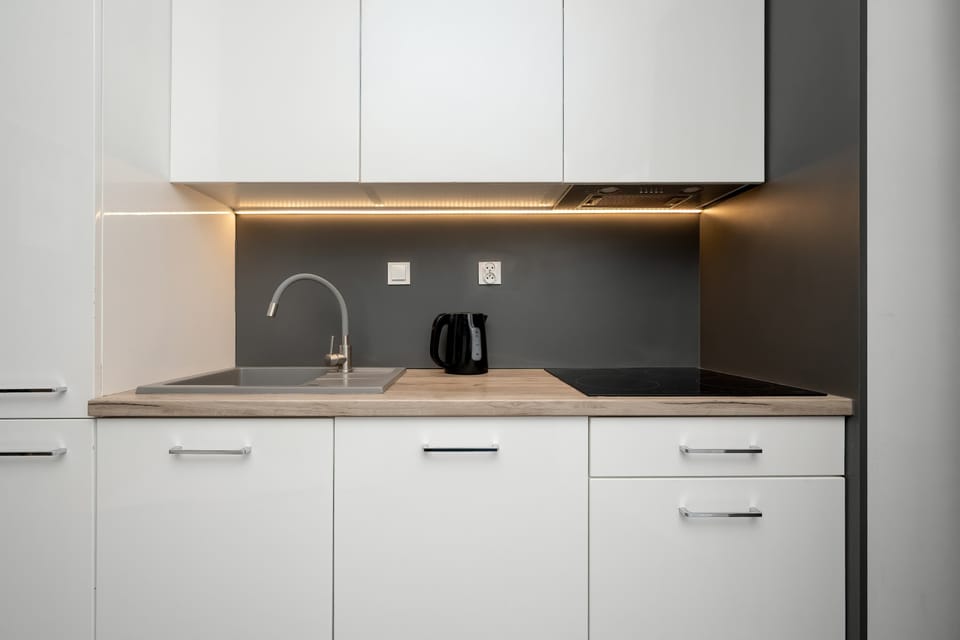 A minimalist kitchenette area displays sleek white cabinetry and a dark grey backsplash with integrated LED lighting. The induction cooktop and modern sink create a clean, functional workspace.
