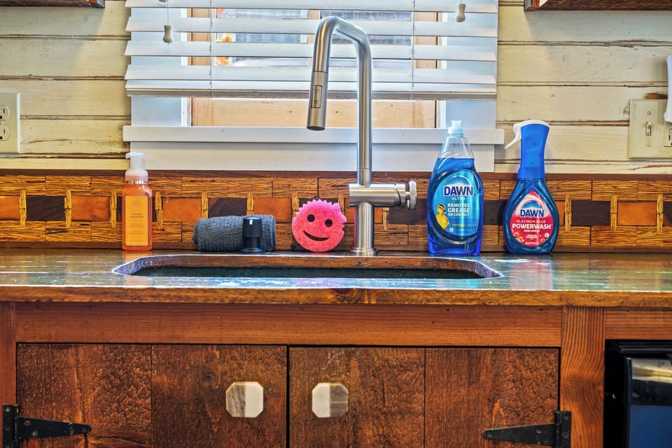 Preparing meals in the ship's galley. An upgraded high-arc faucet matches the washroom hardware, standing over a copper basin sink fully stocked with cleaning supplies and a smiling sponge beneath the window blinds.