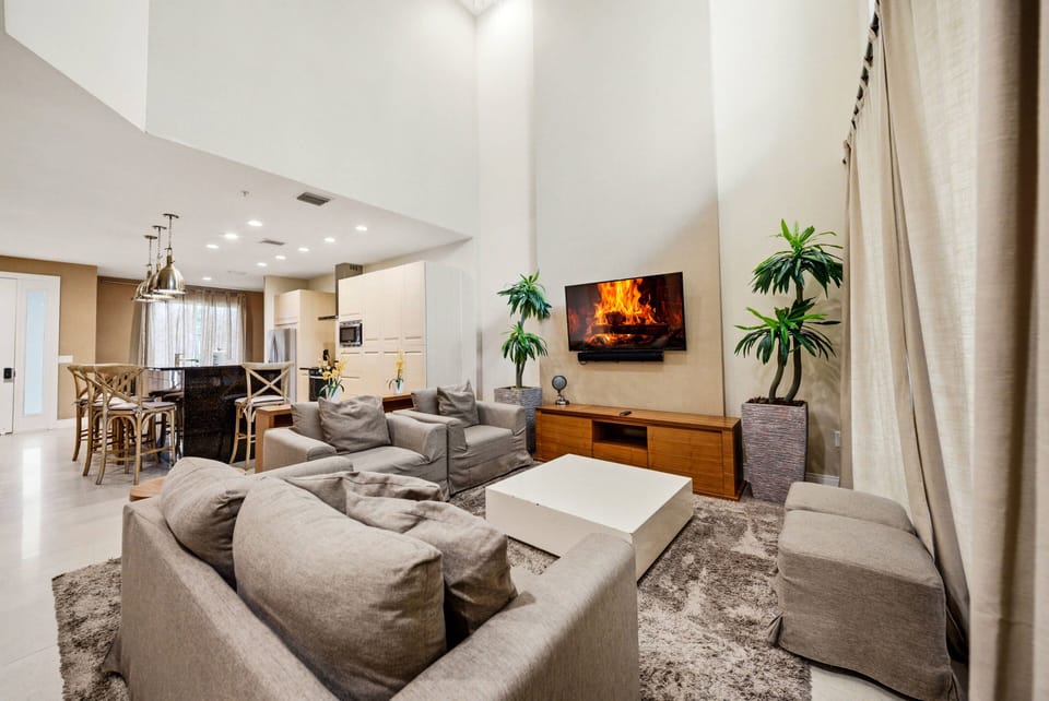 Experience a bright living area with soft neutral tones and comfy seating that invites you to relax. Large windows fill the space with natural light, and a cozy fireplace adds to the stylish decor for a warm, welcoming atmosphere.