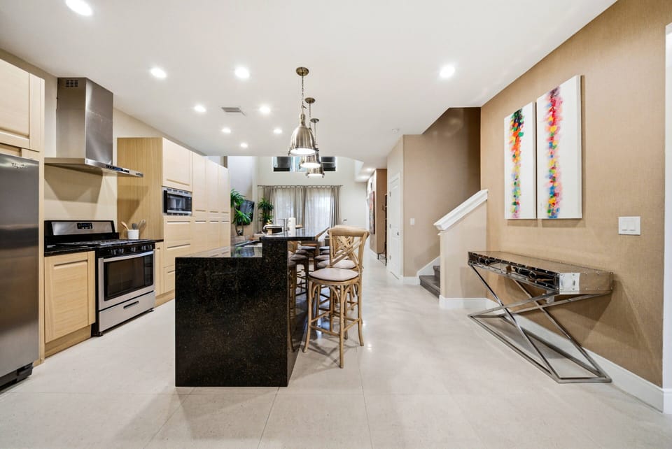 This bright and spacious kitchen boasts sleek cabinetry and modern appliances, highlighted by a striking black island. Elegant pendant lights illuminate the space, and colorful artwork adds personality to the warm-toned walls.