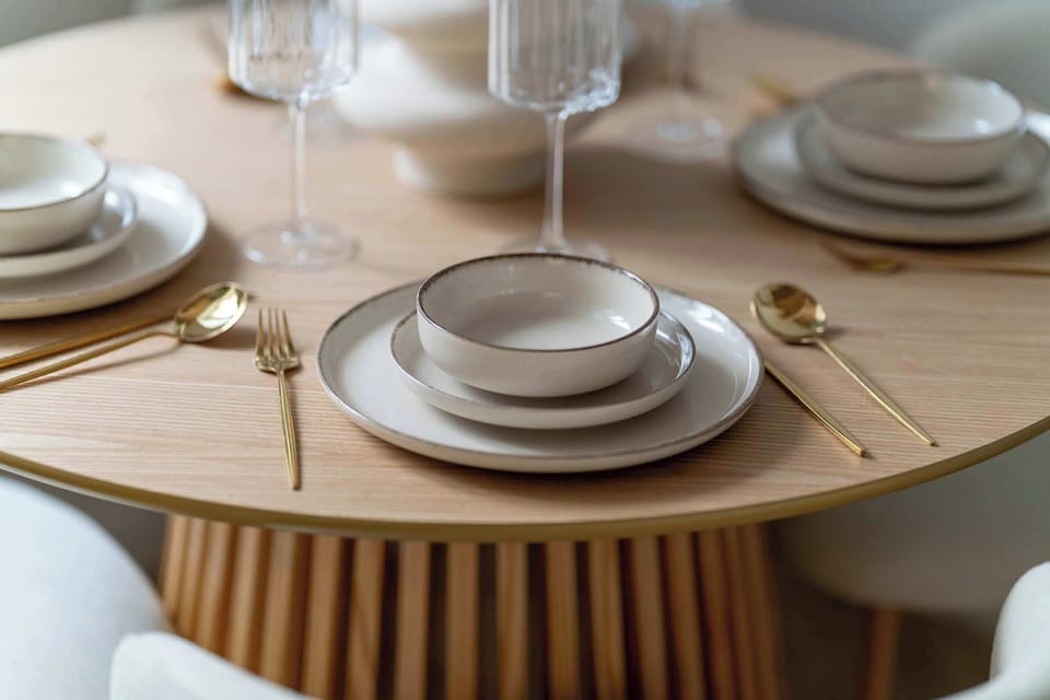 A top-down view of the dining table highlights a sophisticated setting with white stoneware and gold-toned cutlery. The slatted wooden texture of the table serves as a warm, organic base for the minimalist dinnerware.