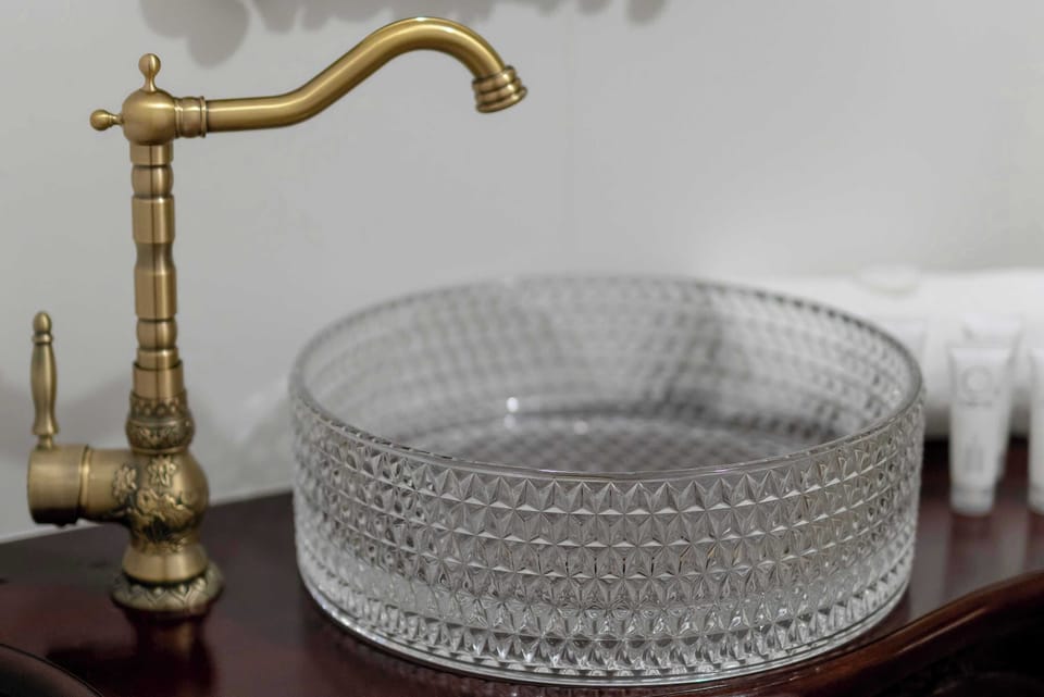 A detailed shot of the bathroom vanity emphasizes the interplay between the textured glass sink and the antique brass hardware. The dark wood of the washstand provides a heavy, grounded contrast to the light reflecting off the glass.