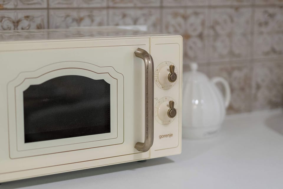 A cream-colored retro microwave with gold accents sits on a kitchen counter against a backdrop of ornate tiles. The appliance’s soft curves and vintage dials complement the apartment's blend of old-world and modern styles.