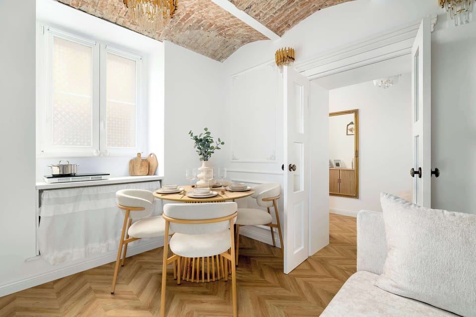 Sunlight streams through a large window onto a round wooden dining table set for four. The view through an open doorway reveals a glimpse of the bedroom, emphasizing the apartment's open and airy layout.