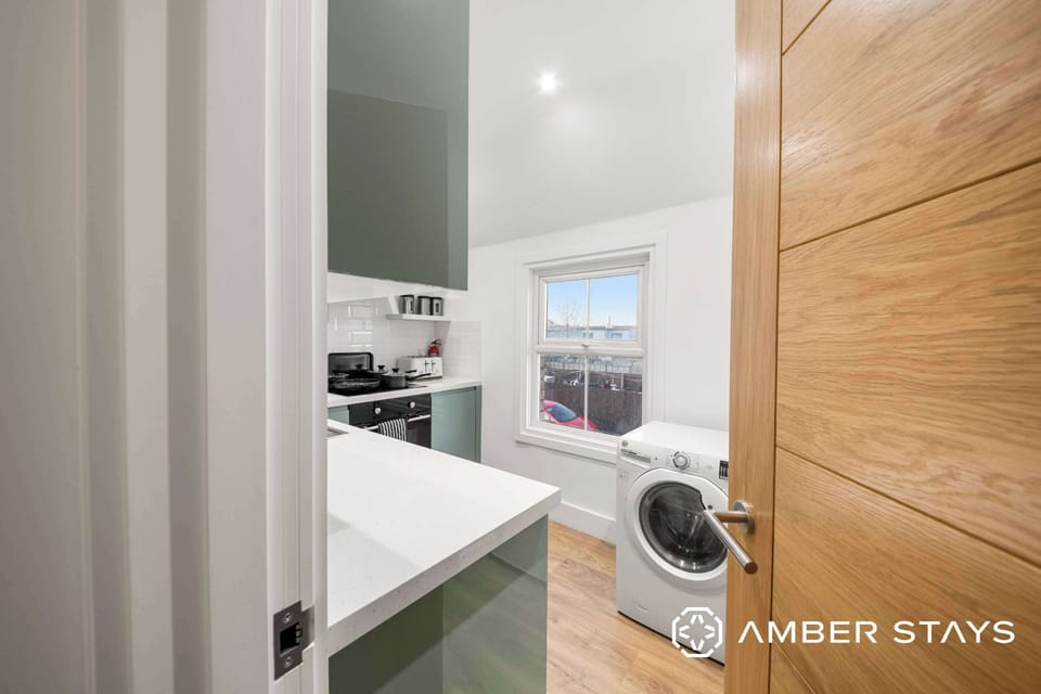 A kitchen viewed through a doorway features green cabinets, white countertops, a washing machine, and a small window in the background.