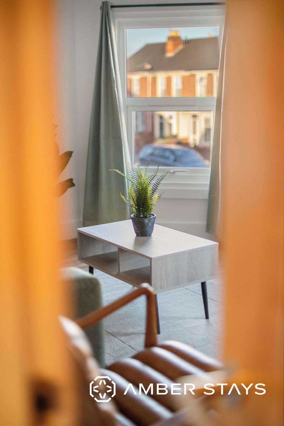 A living room view through a doorway shows a wooden coffee table with a potted plant, a leather armchair in the foreground, and a window with curtains.