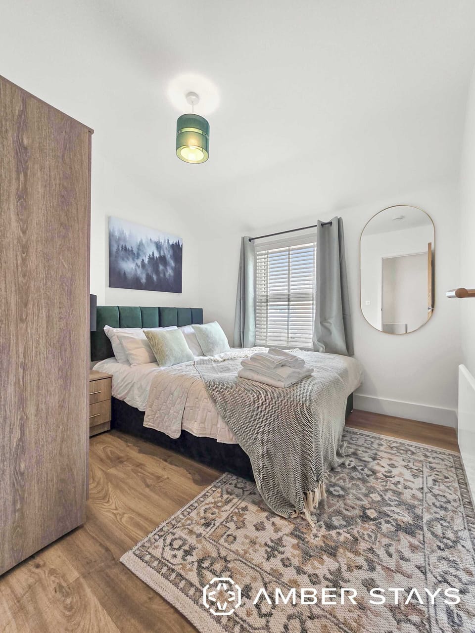 This bright, modern bedroom features a plush double bed, white walls, and light wood floors. Stylish accents include a patterned rug, a large oval mirror, and a green pendant light.