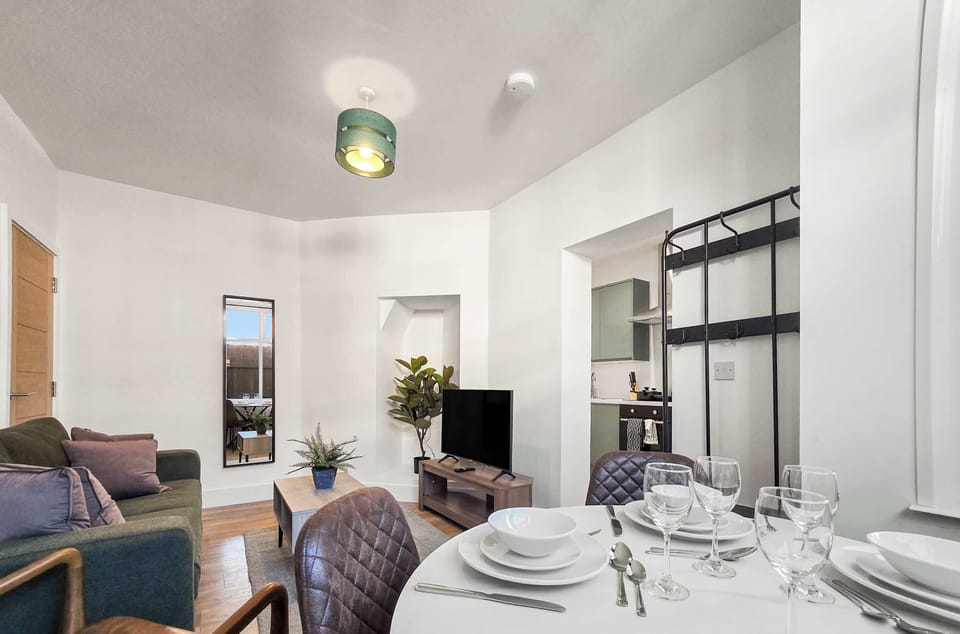 A bright, modern open-plan living and dining space featuring a set table in the foreground. A cozy seating area and small kitchen are visible in the background.