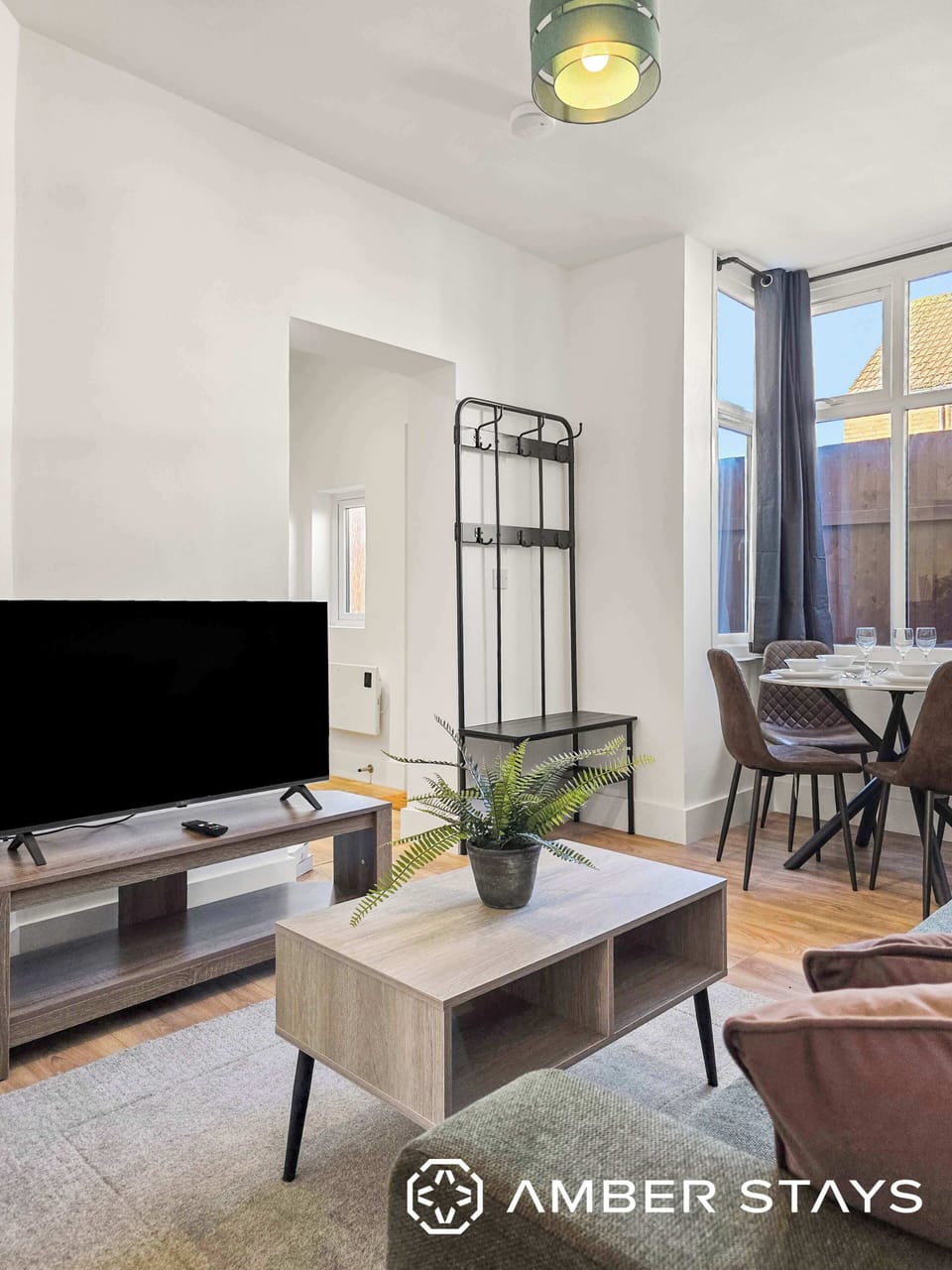  A brightly lit, modern living area featuring light wood furniture and a minimalist black coat rack. A large window with grey curtains illuminates a small dining set in the background.