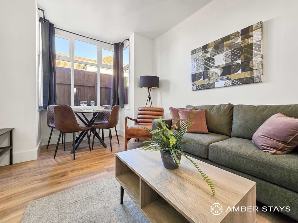 This living room features a modern aesthetic with a green sofa, a light wood coffee table, and a small dining area by the window. Earthy tones and natural light create a warm, inviting atmosphere in the space.