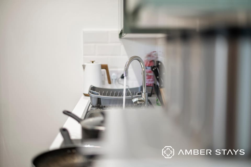 A kitchen counter features a silver faucet, a drying rack, and a white electric kettle against a white tiled backsplash.