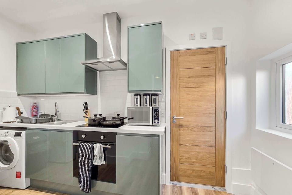A kitchen features grey cabinets, an integrated oven and stovetop with a stainless steel range hood, a washing machine, and a light wood door.