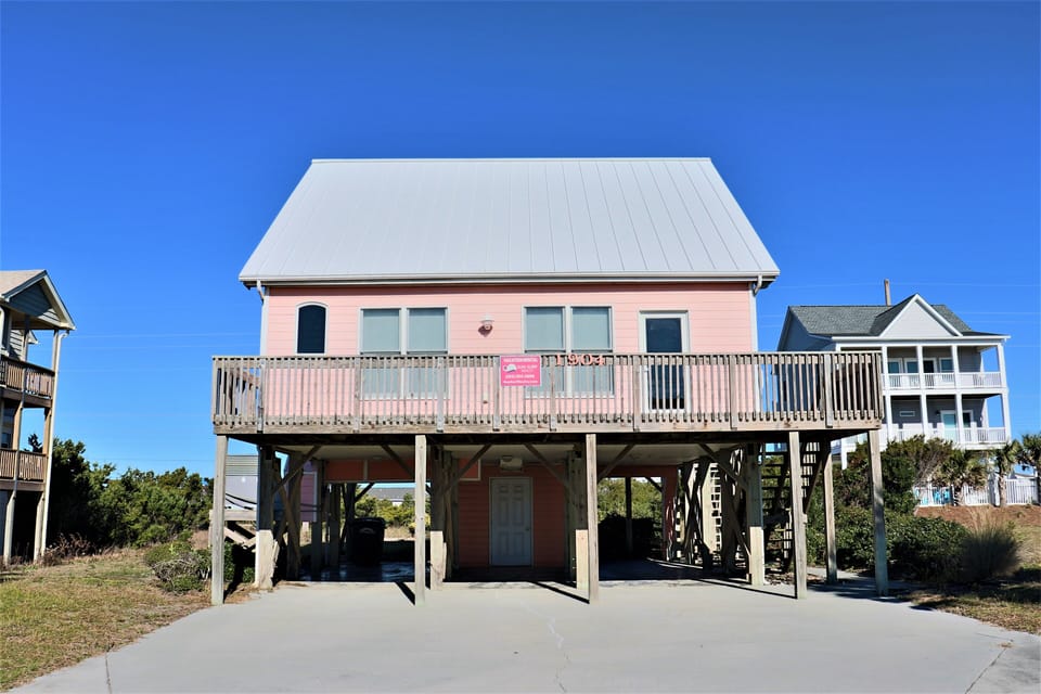 Charming pink beach house on stilts with covered parking and deck, nestled among coastal homes under clear blue skies.