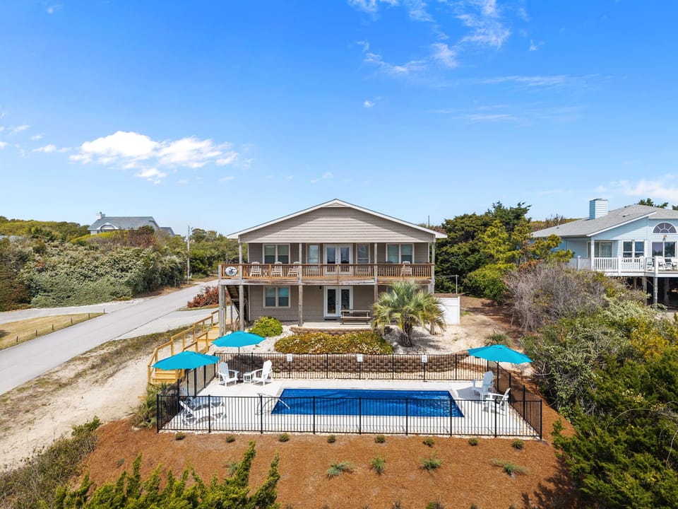 Beachside vacation home features private pool with safety fence, spacious deck with blue umbrellas, and lush coastal landscaping.