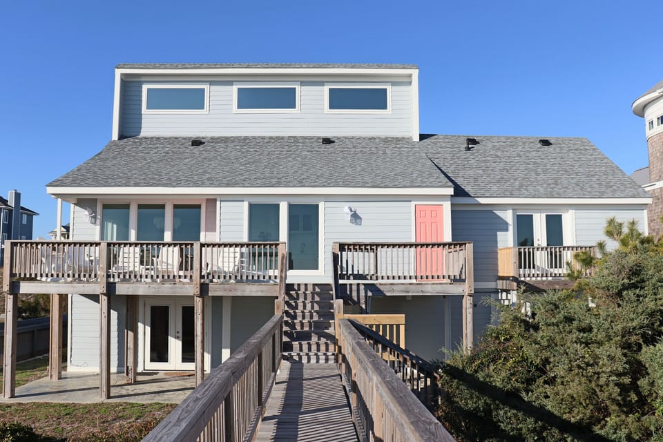 Coastal beach house featuring elevated design with wraparound decks and boardwalk access under clear blue skies.
