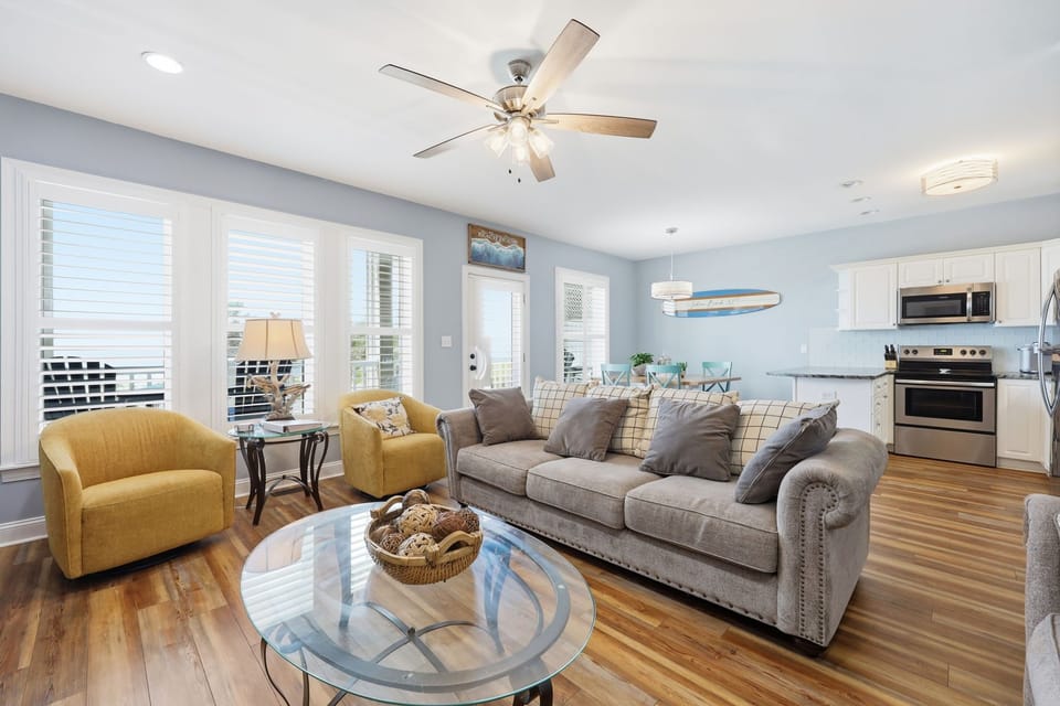 Sink into plush seating in your bright, open-concept living space where coastal breezes flow through shuttered windows and soft blues invite instant relaxation.