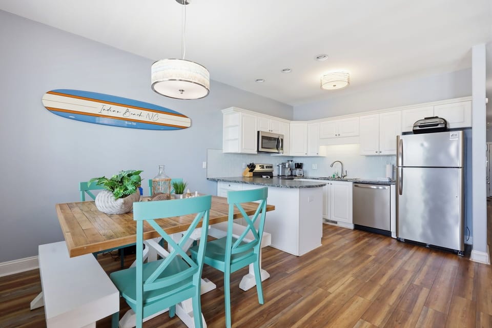 Your open kitchen and dining space blends coastal charm with modern convenience, featuring stainless appliances, a stylish surfboard accent, and turquoise seating that invites relaxed beachside living.