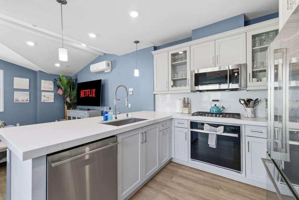 Fully equipped kitchen with stainless steel appliances, gas range, dishwasher, quartz countertops, and open sightlines to the living room and Smart TV