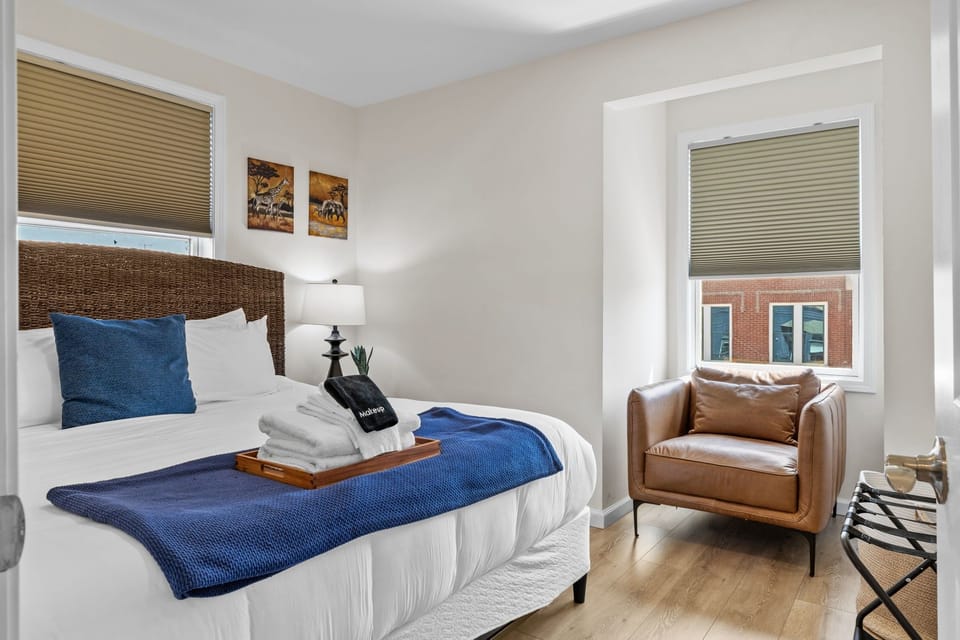 King bedroom with a woven headboard, blue accents, lounge chair, luggage rack, and natural light.