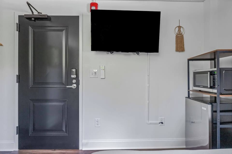A bright, minimalist entry area featuring a sleek black door, wall-mounted TV, and compact kitchenette with a microwave and mini-fridge. Perfect for a convenient, streamlined stay.