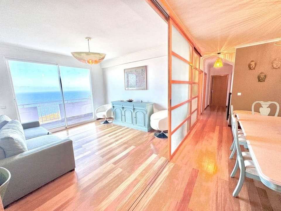A slim, impeccably clean living space focused on the Atlantic horizon. It features a three-seater sofa, two chairs, and a curated collection of Balinese and Peruvian art. Sliding screens open to the sea, creating a light, airy atmosphere.