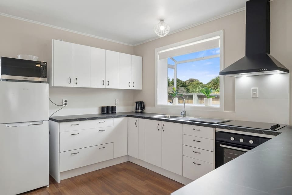 Bright white kitchen with electric cooktop, oven, black rangehood, and garden window view — fully equipped NZ holiday home Rotorua