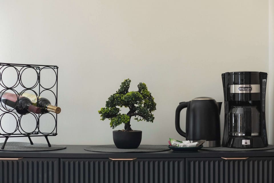 A long black dresser serves as a coffee station, holding a kettle, a drip coffee maker, and a small bonsai tree. A geometric wine rack sits on the left side, adding a decorative yet functional element.