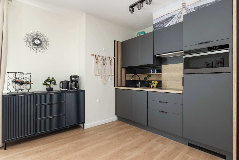 A side view of the kitchenette highlights the contrast between the dark grey units and the light wood textures. The layout is compact but well-equipped with modern appliances.