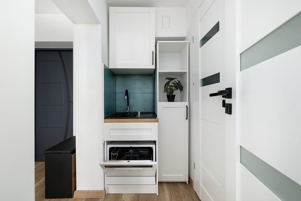 A compact, modern kitchenette designed with clean white cabinetry and subtle gray accents creates a bright and minimalist look. The teal backsplash adds a fresh pop of color, while built-in appliances keep everything sleek and organized.