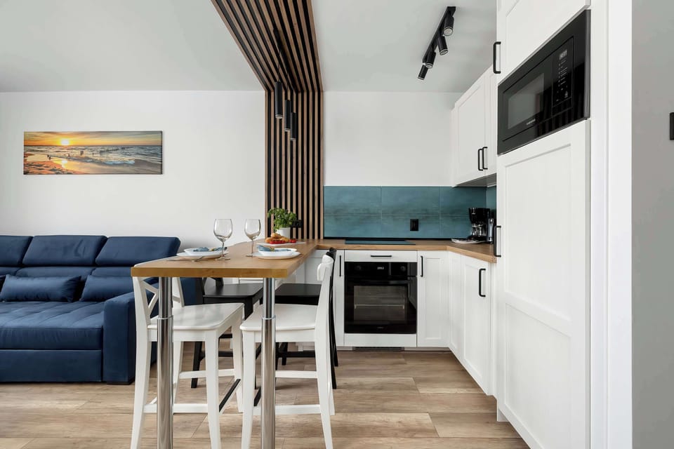 A stylish living area connected to the kitchenette features a comfortable blue sofa and a compact dining table. The space feels airy thanks to large windows and light wood flooring. Subtle decor elements add warmth without clutter.