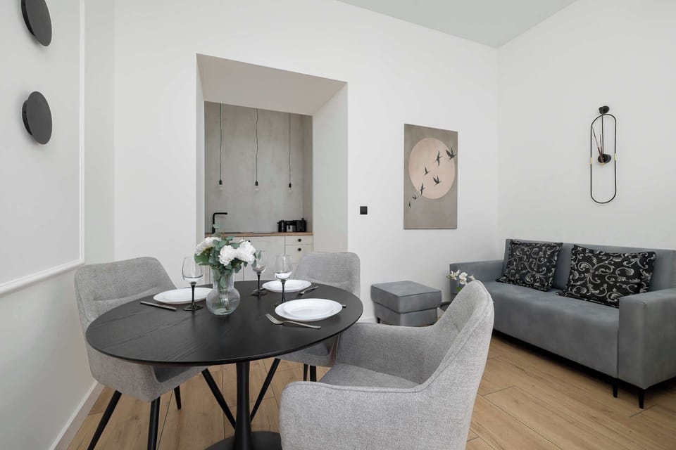 A round, black dining table with three elegant chairs, positioned in the sunniest spot of the apartment. In the background, you can see a gray sofa and minimalist wall decorations in the form of prints.