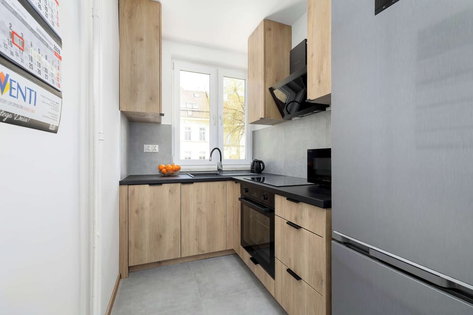 The kitchenette is viewed from an angle that reveals a stainless steel refrigerator and dark appliances. Light wood cabinets contrast beautifully with the dark backsplash and countertop.