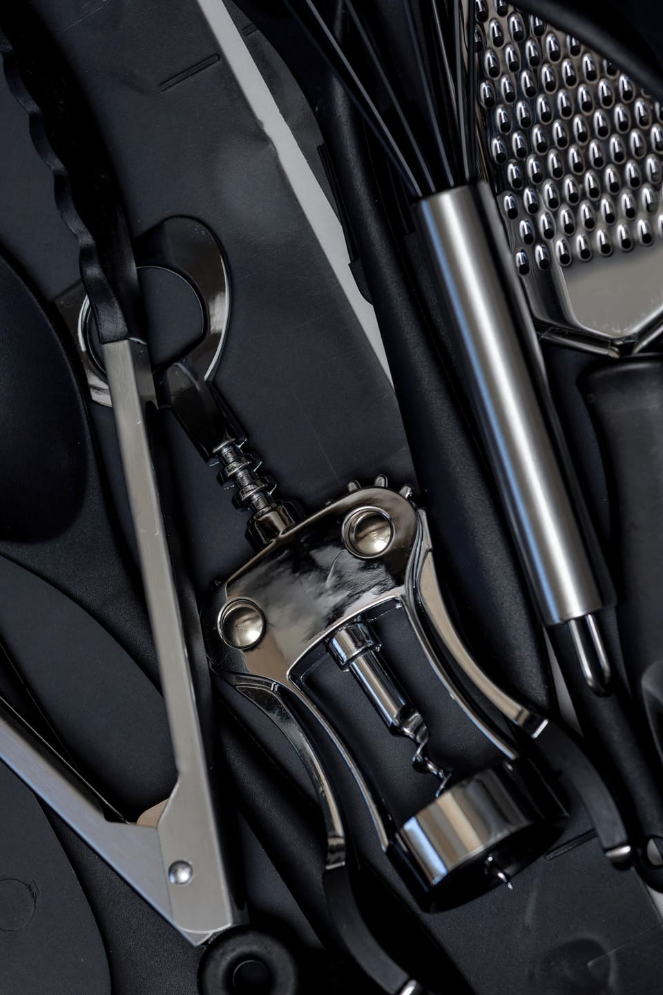 A collection of dark kitchen utensils and a corkscrew are arranged artistically in a drawer or tray. The overhead lighting creates sharp highlights on the metallic and black surfaces.