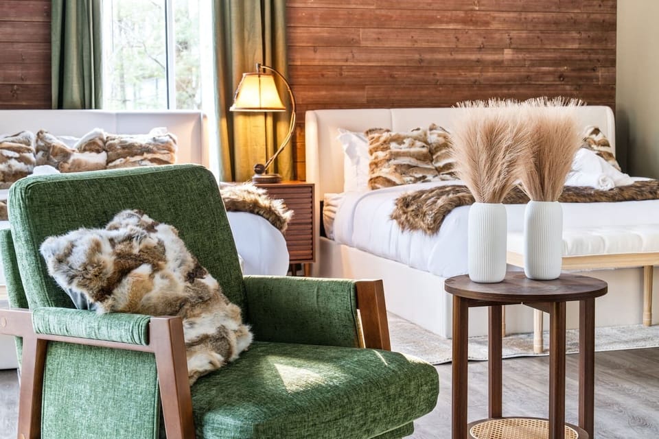 Cozy, lodge inspired bedroom with two beds, warm wood accents, and a unique antler chandelier. French doors open to the deck, with a seating area perfect for relaxing and enjoying peaceful forest views.