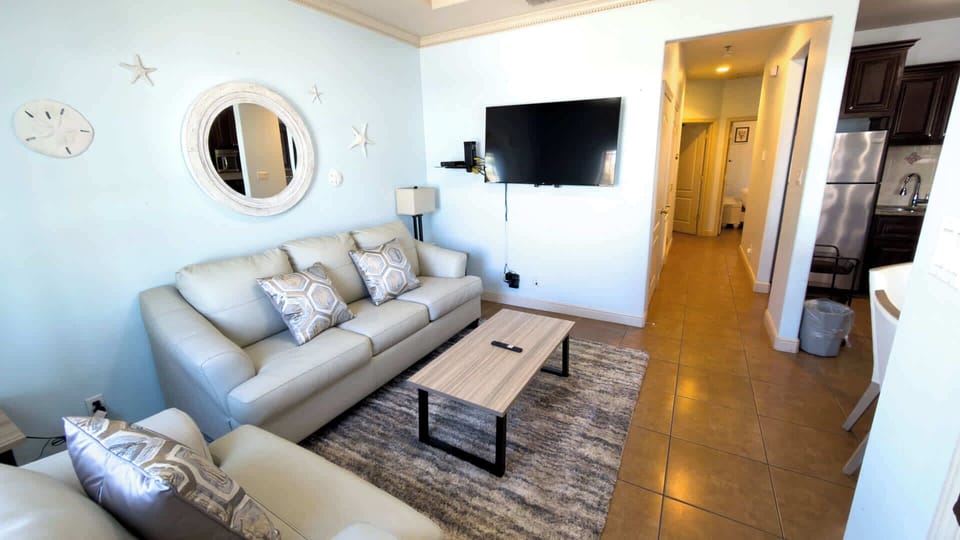 Bright, modern living space featuring cozy sofas, a sleek coffee table, and a stylish accent mirror. Enjoy an open layout flowing into the kitchen, soft lighting, and a welcoming setup ideal for relaxing, streaming, or gathering after a day out.