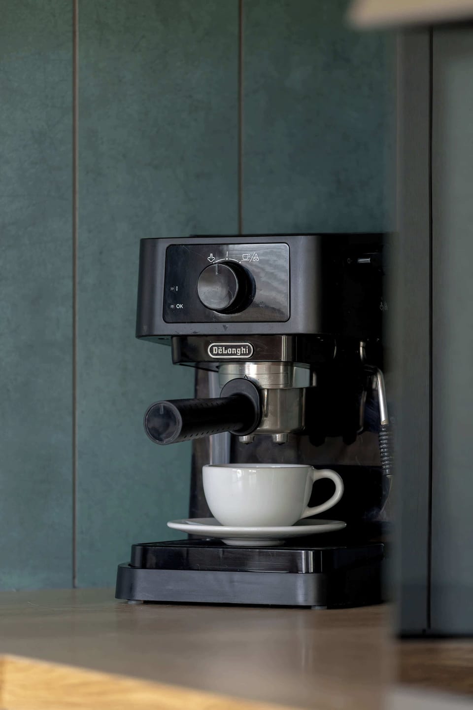 A culinary detail shot highlights a professional black espresso machine sitting on a light wood countertop. The dark teal tiles in the background provide a rich color contrast.