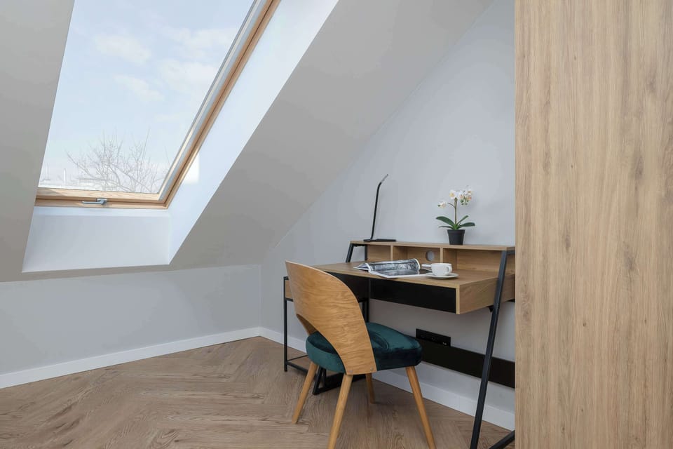 A compact office nook includes a stylish wooden desk and a green velvet chair. A large window provides a view and plenty of daylight for the workspace.
