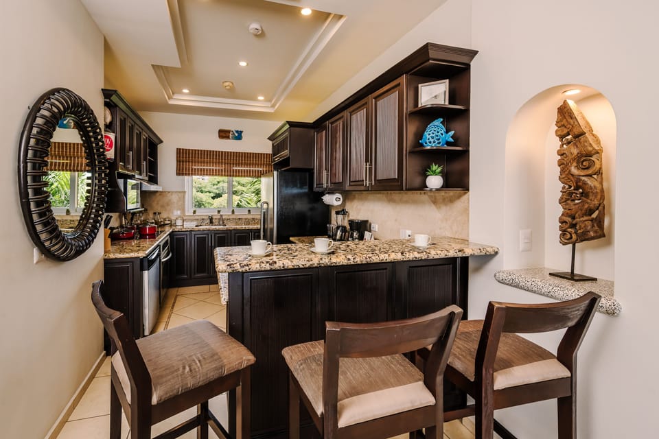 Kitchen breakfast bar with three upholstered stools, granite counter, dark cabinetry, round rattan mirror, and tropical wood sculpture.