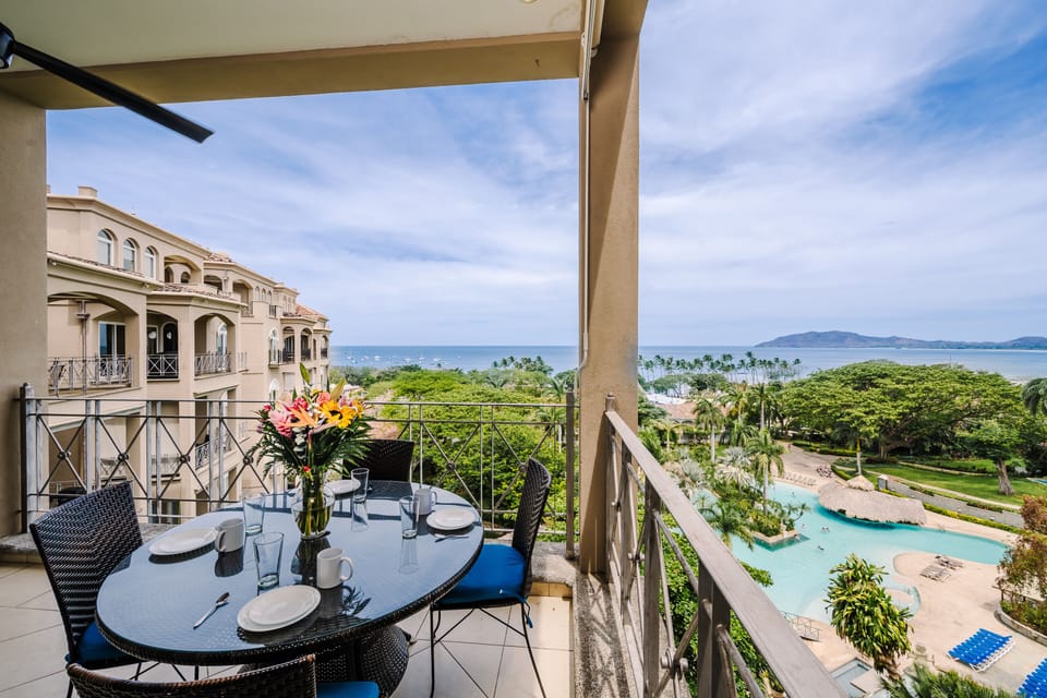 Balcony dining table with fresh flowers overlooking a free-form resort pool and Tamarindo Bay beyond.