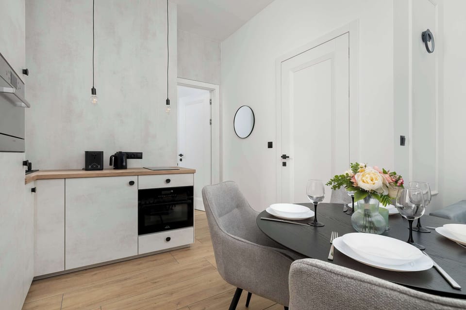 A functional kitchenette with white cabinet fronts, a black oven, and a light wood-textured countertop, connecting to the hallway leading to the bedroom.