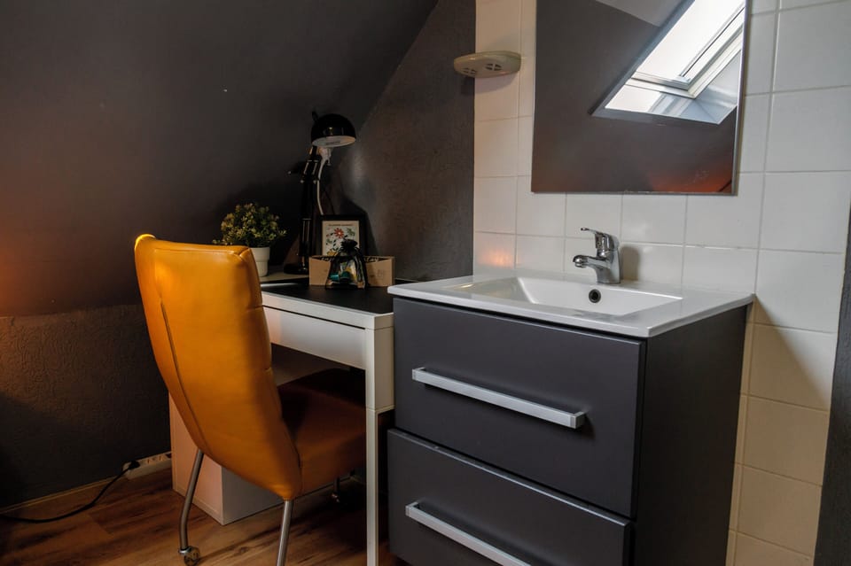 Stylish corner with modern sink, vanity unit, and a cozy workspace featuring a desk, chair, and soft lighting under a sloped ceiling.