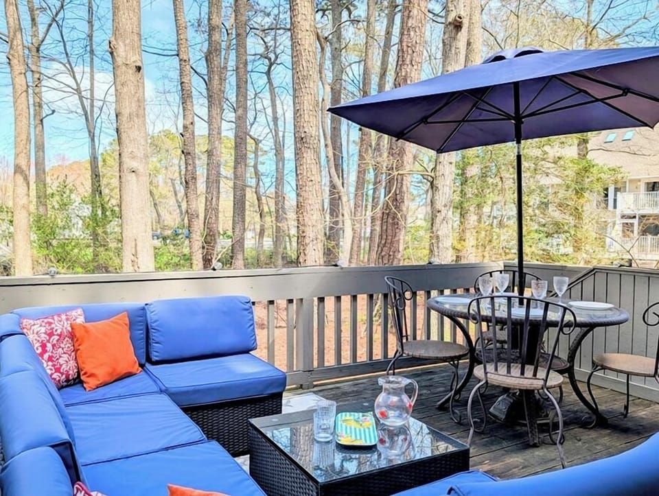 2306 Round Robin Way Sea Colony West Outdoor Wood Deck with Blue - An outdoor wooden deck balcony featuring a blue cushion sectional sofa, a coffee table, and an umbrella-shaded dining table set against a backdrop of mature trees.