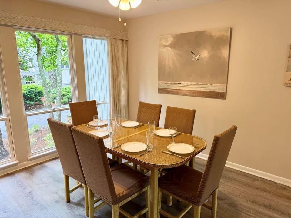 2306 Round Robin Way Sea Colony West Dining Room Glass Top Table - A warm dining space featuring a glass-topped wicker dining table paired with six brown upholstered high-back chairs, wood-look flooring, large bright windows, and a prominent canvas painting of a seagull over the ocean.