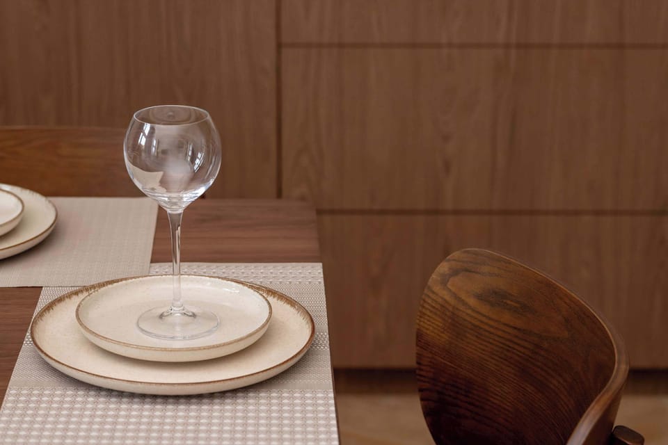 A dining table setting features a ceramic plate and a wine glass on a checkered placemat. The dark wood of the chair in the foreground complements the warm tones of the cabinetry in the background.