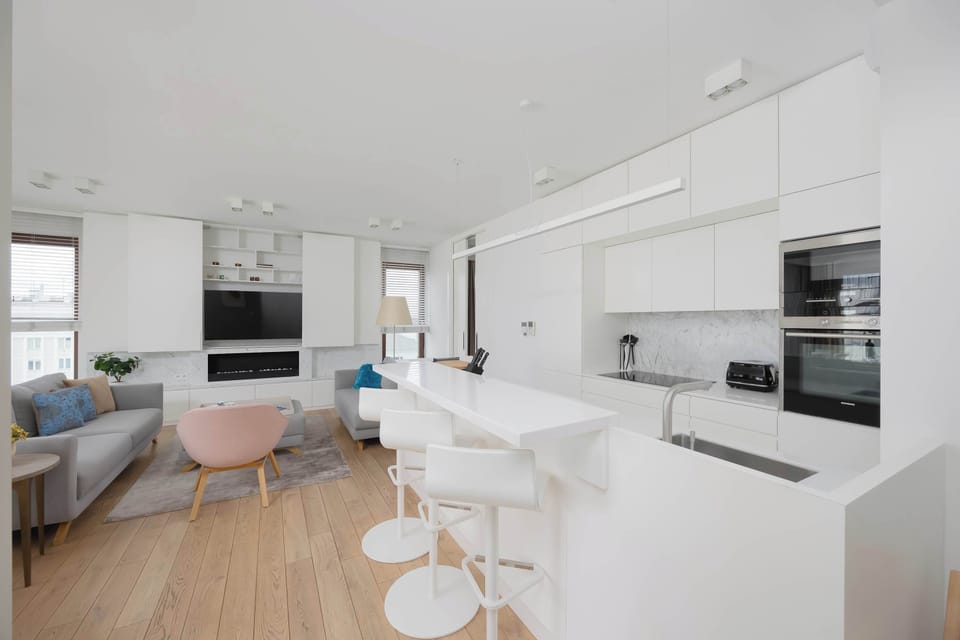 An open-plan living area connects seamlessly with the kitchenette, featuring soft pastel accents and light wooden flooring. The space feels airy and welcoming, ideal for relaxing or socializing.