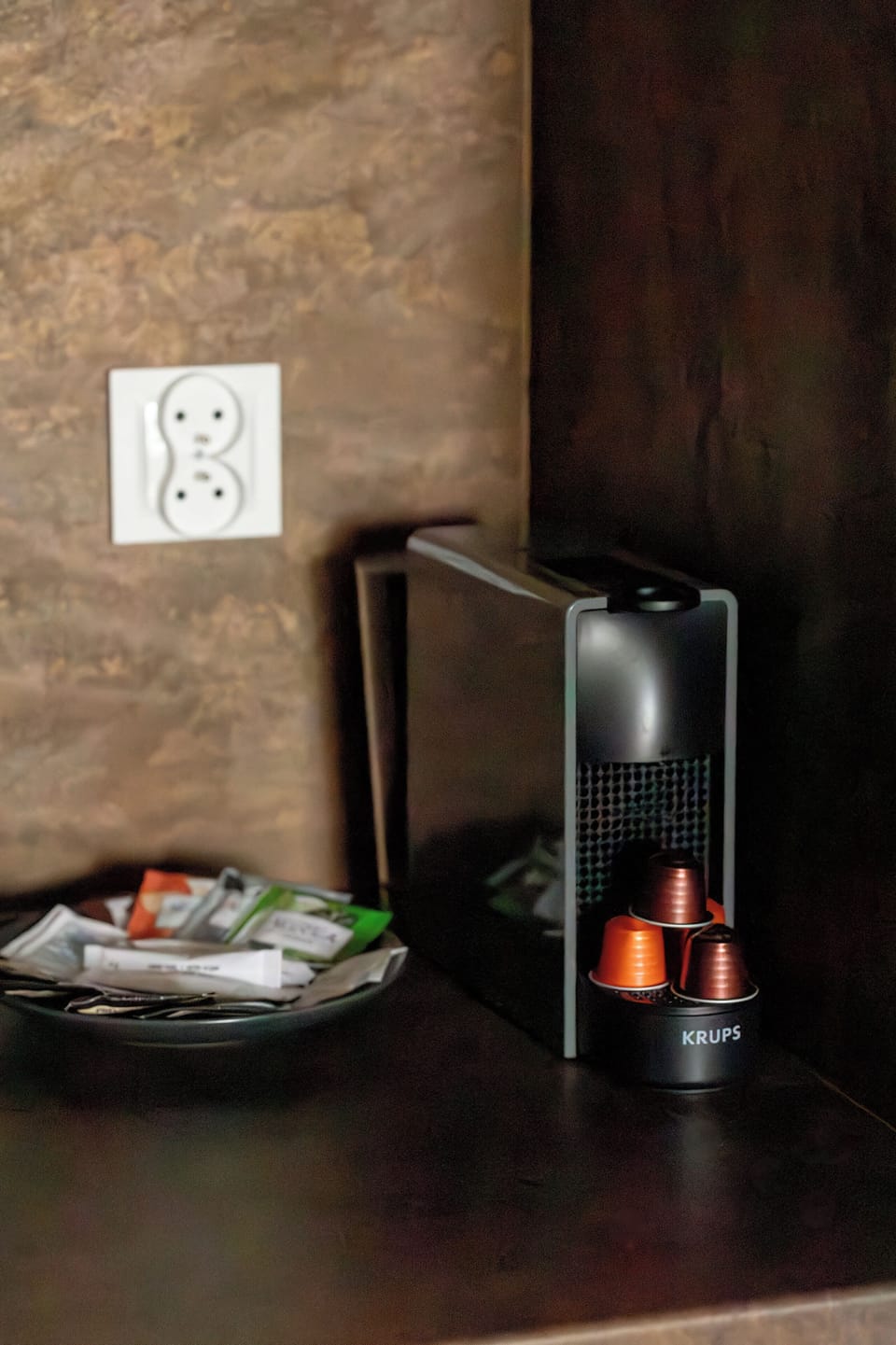 A sleek coffee pod machine sits on a dark countertop beside a wall outlet, with a cup positioned underneath the spout. Next to it, a small tray holds coffee capsules, creating a tidy and modern kitchen corner setup.