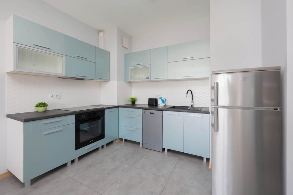 L-shaped kitchen arrangement maximizes storage with sleek, handle-less cabinets in a soft pastel blue. High-end appliances, including a built-in oven and dishwasher, are neatly integrated into the design.