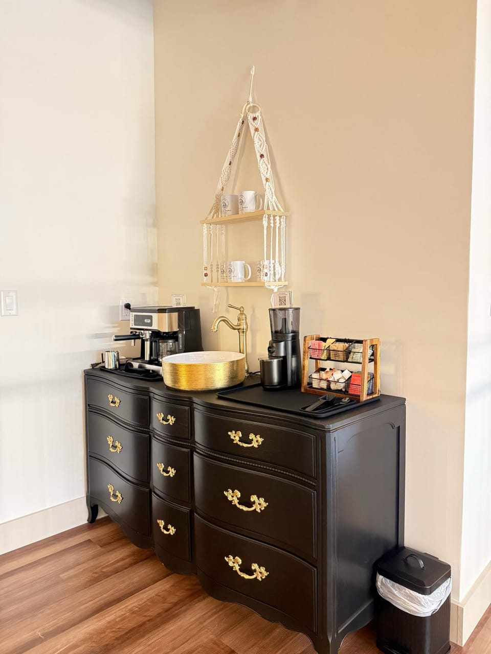 Black vintage-style dresser topped with brass faucet, coffee maker, grinder, and hanging macrame shelf holding mugs.