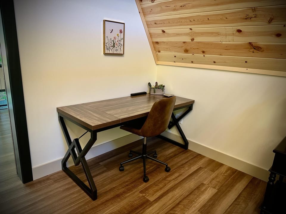 Corner workspace with wood-paneled slanted ceiling, wood-grain desk, brown leather swivel chair, and floral wall art.